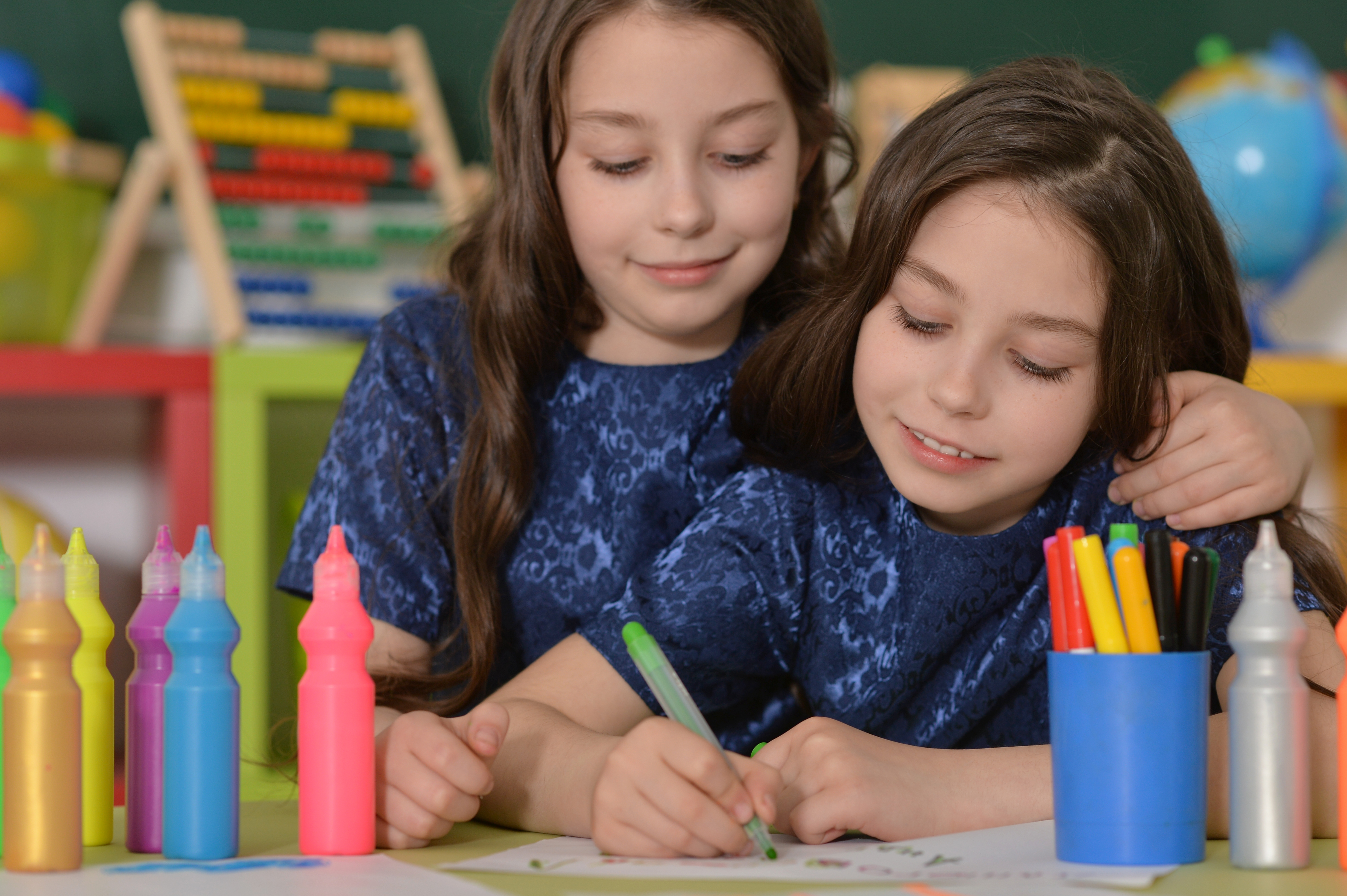 Dwie dziewczynki siedzą przy biurku i piszą kolorowymi mazakami.