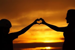 Dwie osoby łączą swoje dłonie tworząc kształt serca na tle zachodu słońca.