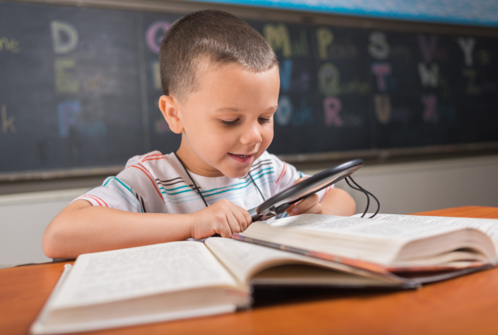 Uśmiechnięty chłopiec uczy się z książki, korzysta z lupy. Jak się uczyć? Styl uczenia się - wzrokowiec